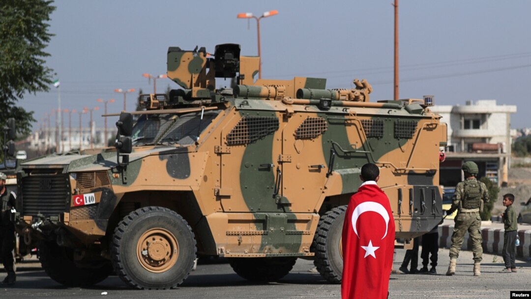 FILE - A boy, with a Turkish flag draped over his shoulders, stands in front of a Turkish military vehicle in the town of Tal Abyad, Syria, Oct. 23, 2019.