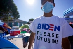 FILE - A Honduran man seeking asylum in the United States wears a shirt that reads, "Biden please let us in," as he stands among tents that line an entrance to the border crossing, March 1, 2021, in Tijuana, Mexico.