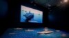 A TV screen showing the U.S. Navy fleet sail in formation near the models of Liaoning aircraft carrier with navy frigates and submarines on display at the military museum in Beijing, Aug. 1, 2019.
