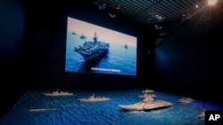 A TV screen showing the U.S. Navy fleet sail in formation near the models of Liaoning aircraft carrier with navy frigates and submarines on display at the military museum in Beijing, Aug. 1, 2019.